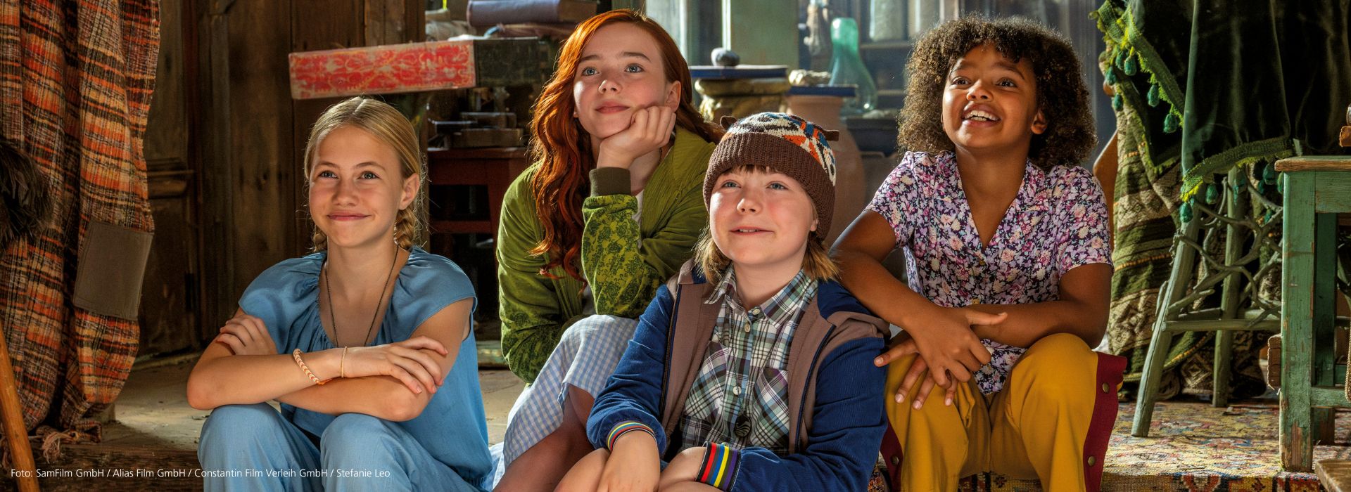 Szene aus dem Film ‘Ein Mädchen namens Willow’: Die vier Hexenfreundinnen Valentina, Willow, Gretchen und Lotti sitzen lächelnd nebeneinander auf einer Treppenstufe, umgeben von bunten Tüchern und Möbeln.